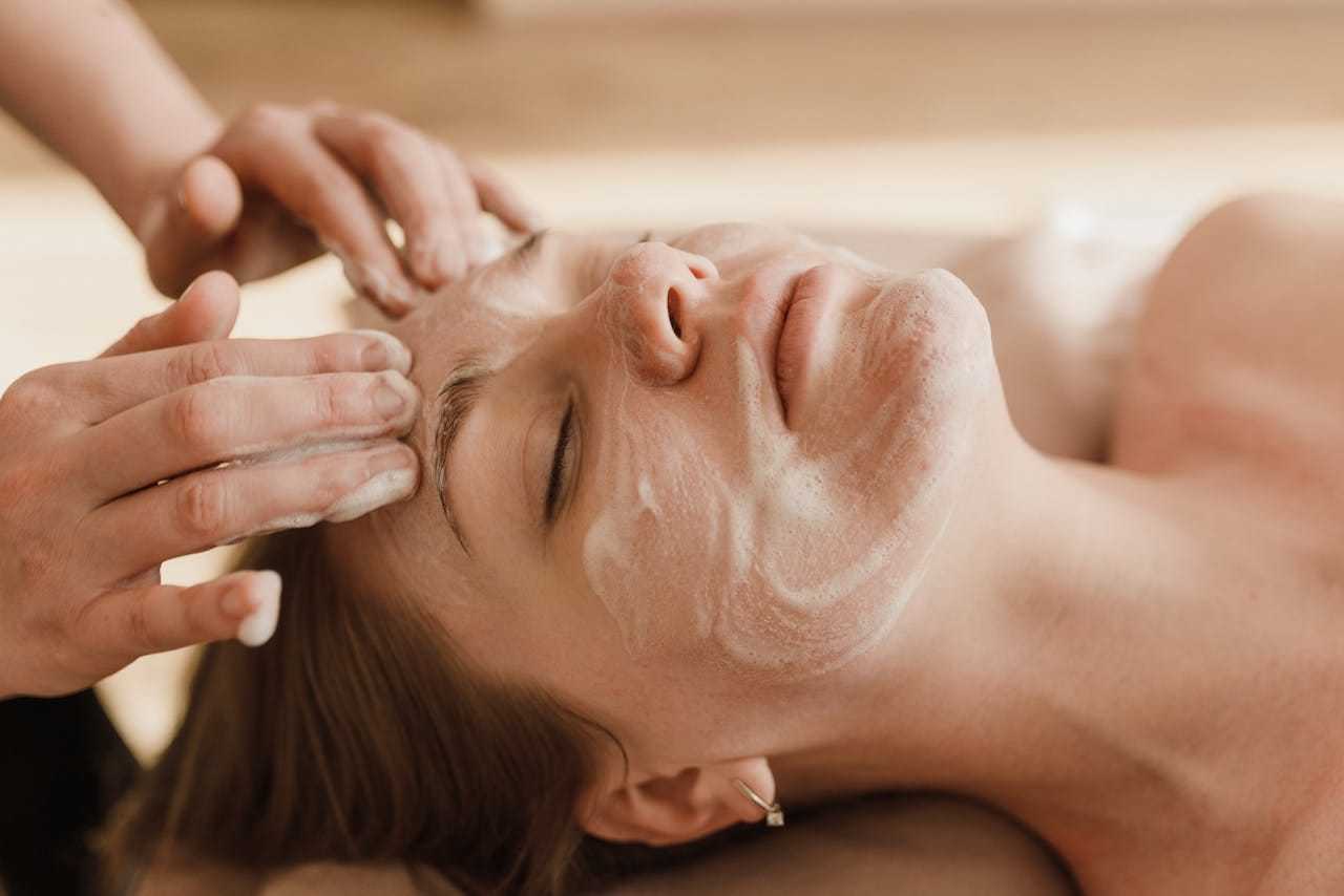 Person receiving a facial treatment, with eyes closed and hands applying a face mask.