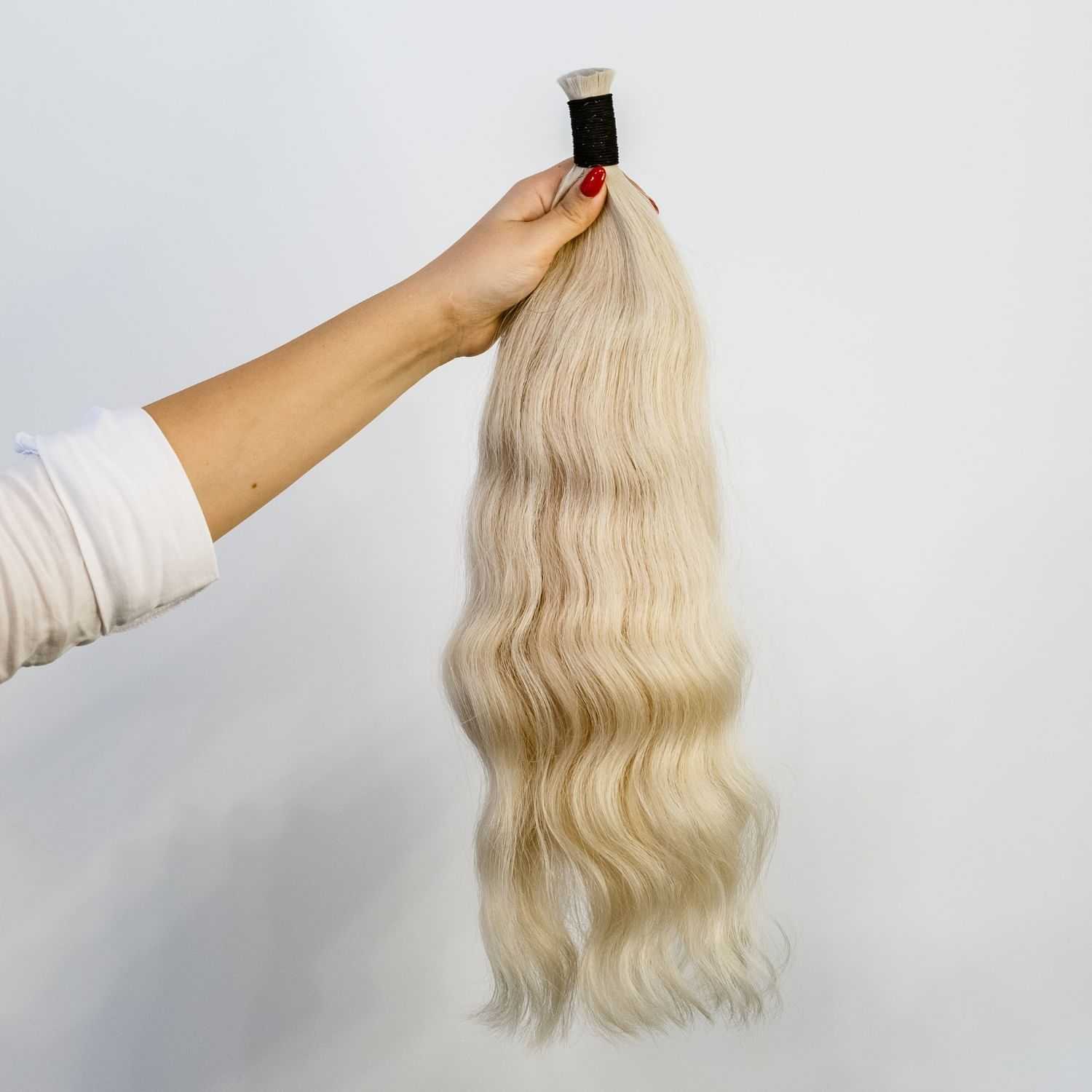 Hand holding a bundle of long, wavy, blonde hair extensions against a plain background.