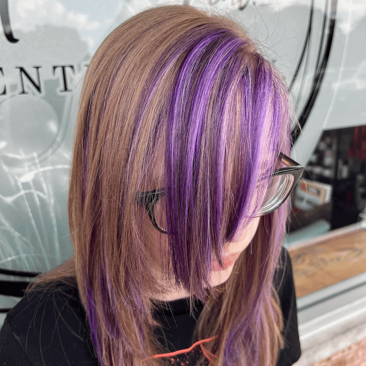 Woman with brown hair and purple highlights standing outside a salon with a sign in the background.