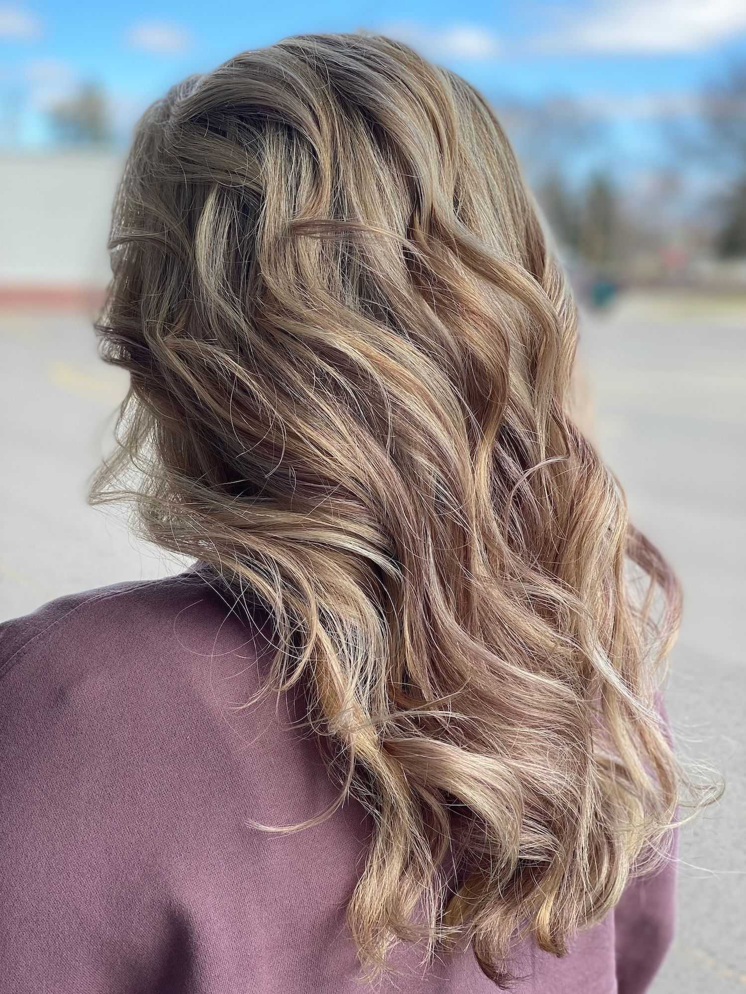 Woman with long, wavy blonde hair wearing a maroon top, photographed from the back outdoors.