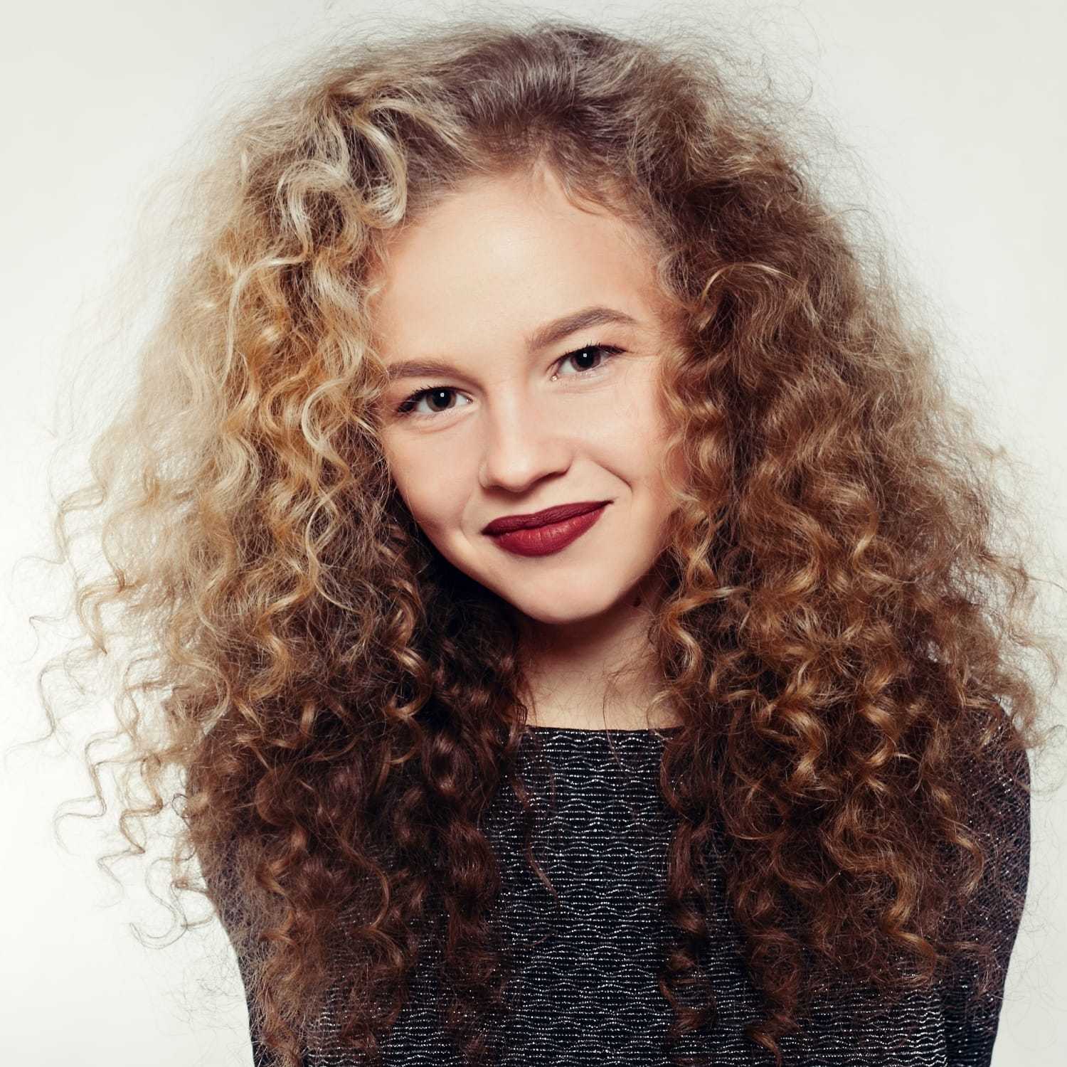 Young woman with curly blonde hair and red lipstick, wearing a dark textured top, smiling.