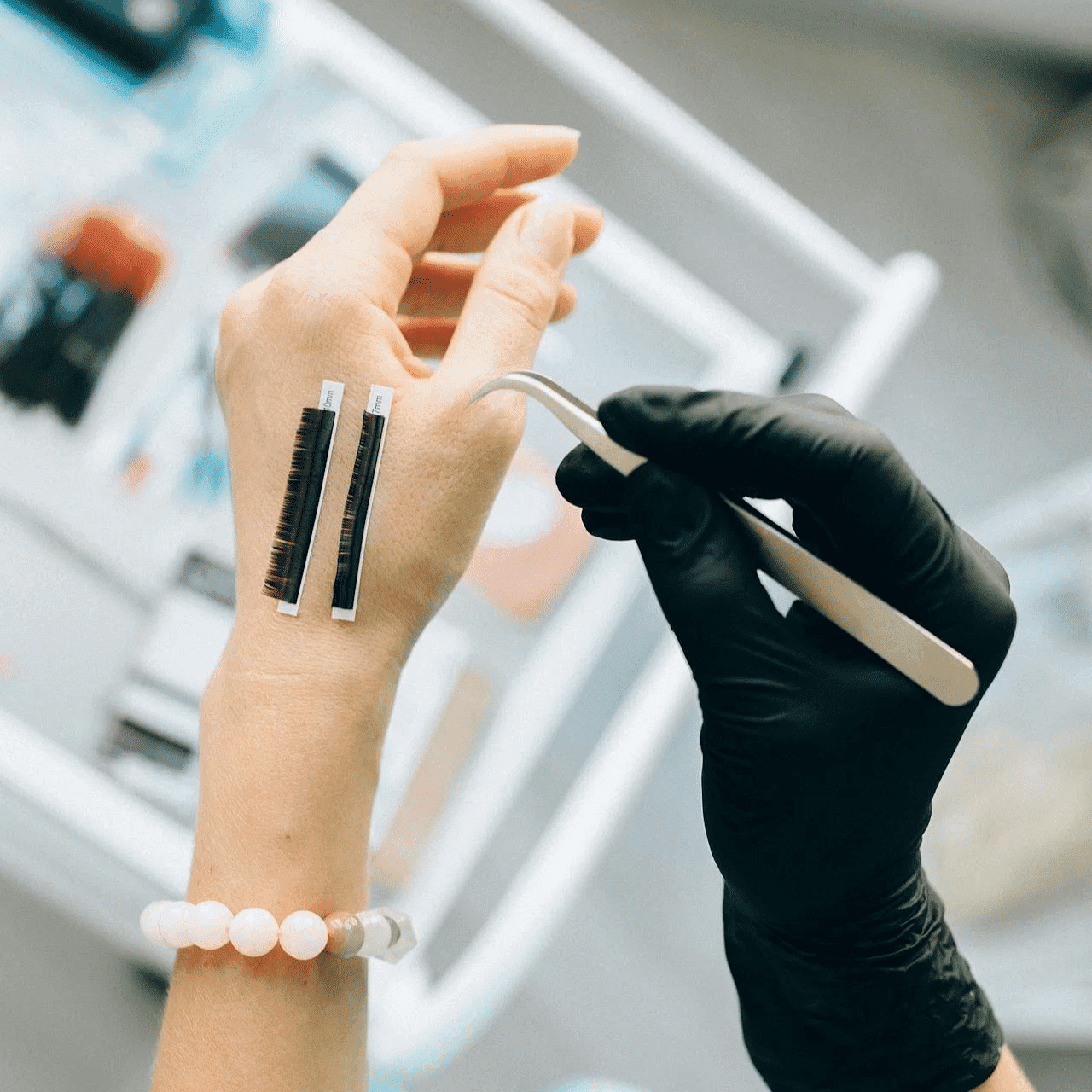 A person with eyelash extension strips on their hand holds tweezers, preparing for application.