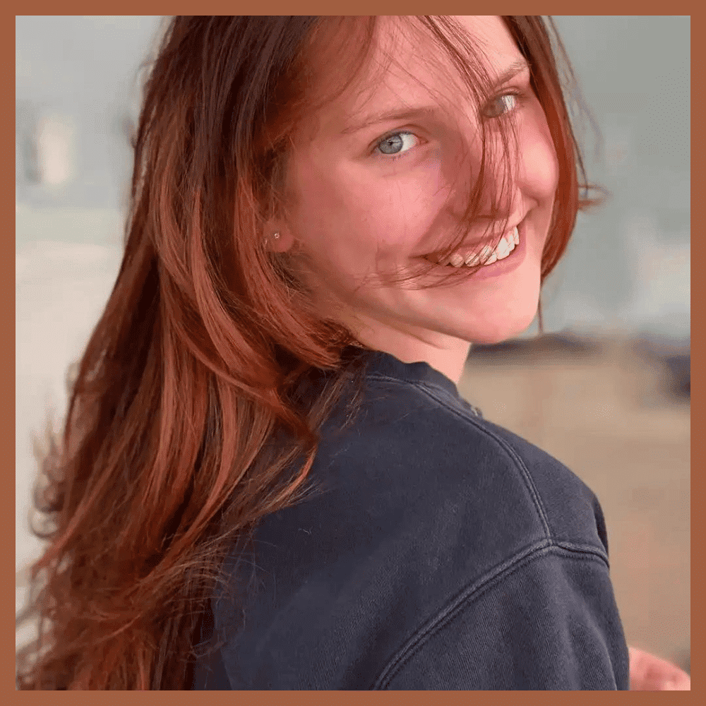 Smiling woman with long brown hair in a blue top, with wind blowing her hair and soft background.